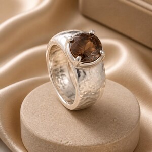 May include: A silver ring with a large, oval, brown gemstone. The ring has a textured band and is set on a neutral-colored, round pedestal. The background features a soft, draped fabric.