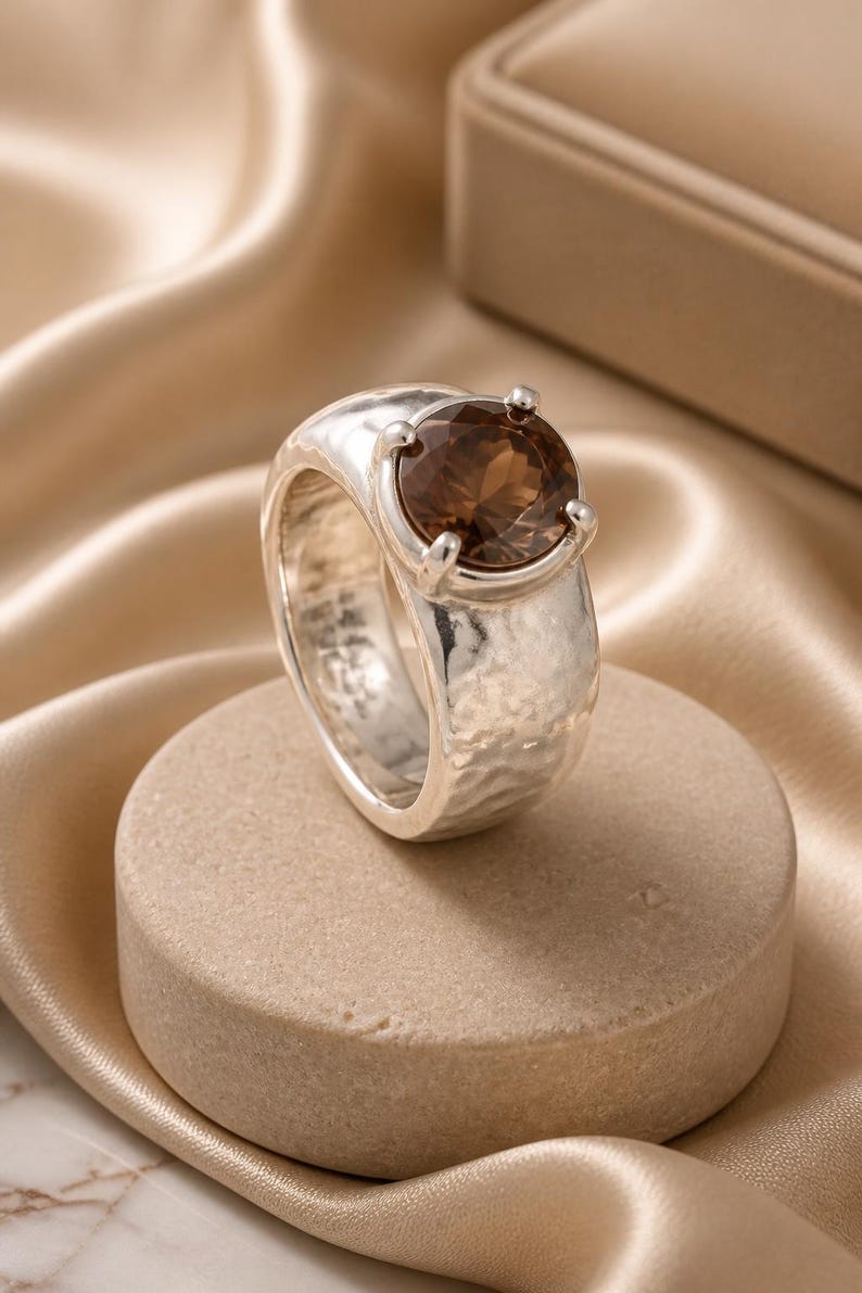 May include: A silver ring with a large, oval, brown gemstone. The ring has a textured band and is set on a neutral-colored, round pedestal. The background features a soft, draped fabric.