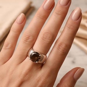May include: A silver ring with a large, round, smoky quartz gemstone. The ring has a wide band with a textured surface. The gemstone is a deep brown color and is set in a simple, classic style. The ring is on a hand with polished nails.