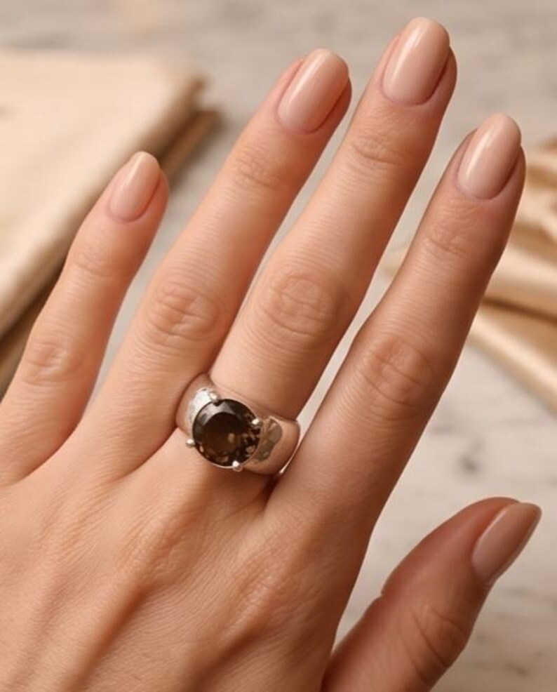 May include: A silver ring with a large, round, smoky quartz gemstone. The ring has a wide band with a textured surface. The gemstone is a deep brown color and is set in a simple, classic style. The ring is on a hand with polished nails.
