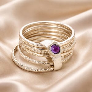 May include: A stack of silver rings featuring a single band with a round, purple amethyst gemstone set in a smooth bezel. The other rings in the set have a textured, hammered finish, creating a layered appearance with varying band widths.