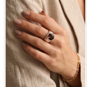 May include: A silver ring with a large, oval, smoky quartz gemstone. The ring is on a hand with neutral-toned nail polish, and a gold chain bracelet is visible. The background is a beige fabric.