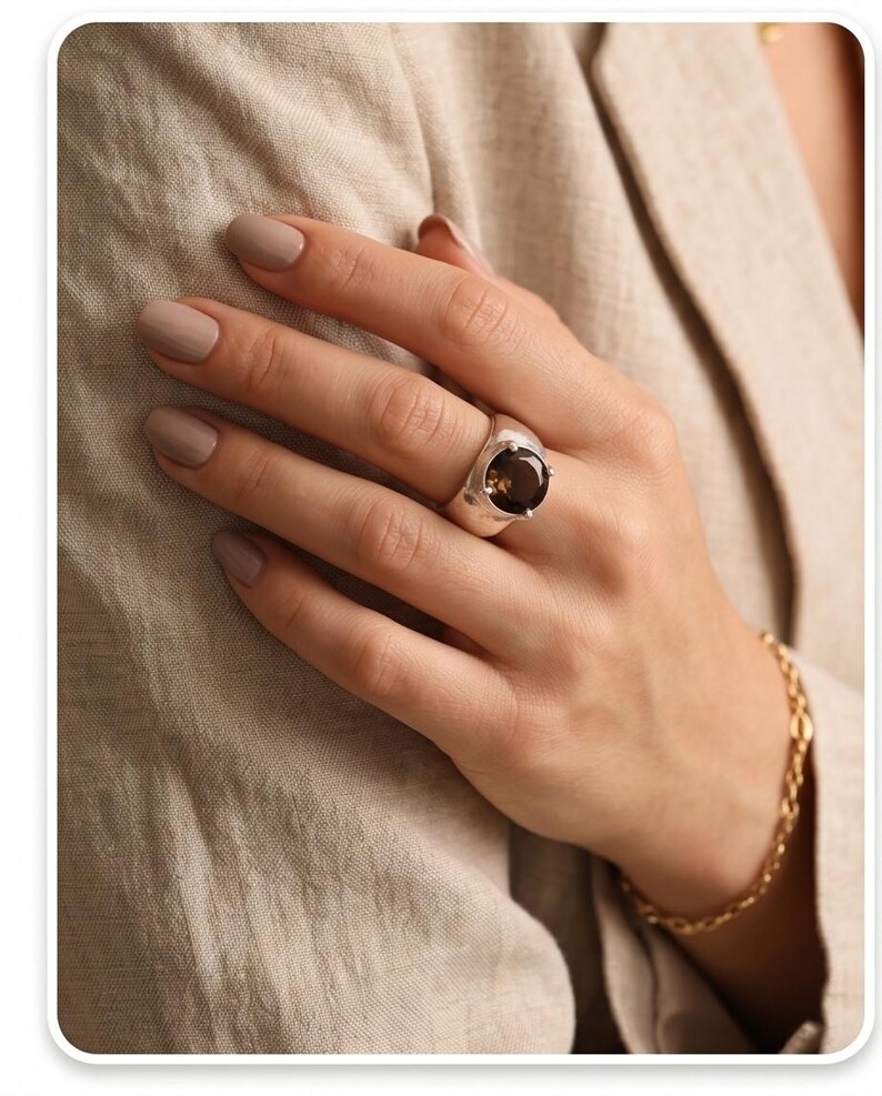 May include: A silver ring with a large, oval, smoky quartz gemstone. The ring is on a hand with neutral-toned nail polish, and a gold chain bracelet is visible. The background is a beige fabric.
