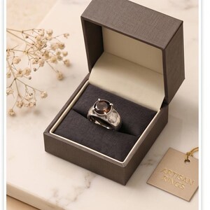May include: A silver ring with a large, round, brown gemstone sits in a dark gray box with a light beige interior. The box is open, revealing the ring. A small tag with the words "ARTISAN RINGS" hangs nearby.