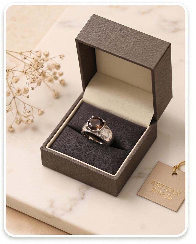 May include: A silver ring with a large, round, brown gemstone sits in a dark gray box with a light beige interior. The box is open, revealing the ring. A small tag with the words "ARTISAN RINGS" hangs nearby.