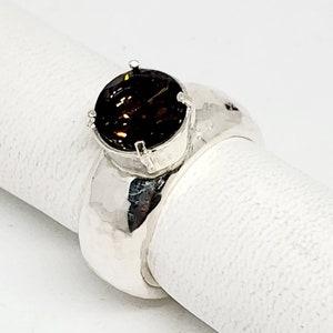 May include: A silver ring with a large, round, dark brown gemstone. The ring has a wide band with a hammered texture. The gemstone is set in a four-prong setting, and the ring is displayed on a white surface.