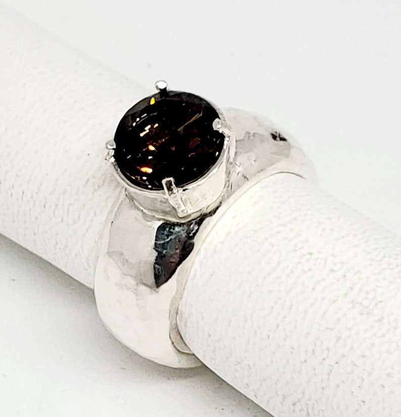 May include: A silver ring with a large, round, dark brown gemstone. The ring has a wide band with a hammered texture. The gemstone is set in a four-prong setting, and the ring is displayed on a white surface.