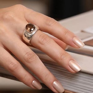 May include: A silver ring with a large, oval-shaped, brown gemstone. The ring has a wide band with a textured design. The ring is on a hand with polished, light-colored nails. The ring is a statement piece.