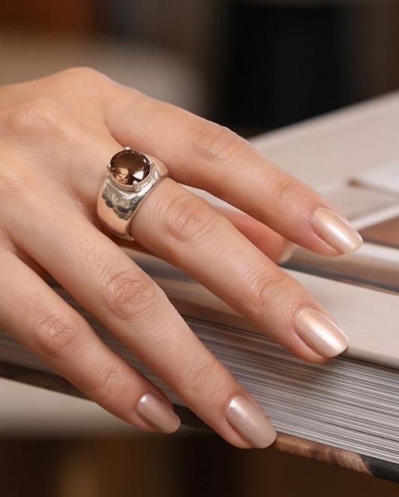 May include: A silver ring with a large, oval-shaped, brown gemstone. The ring has a wide band with a textured design. The ring is on a hand with polished, light-colored nails. The ring is a statement piece.