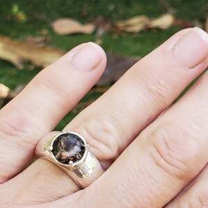 May include: A silver ring with a large, round, smoky quartz gemstone. The ring has a textured band and is set against a blurred green background. The gemstone is a translucent brown color.