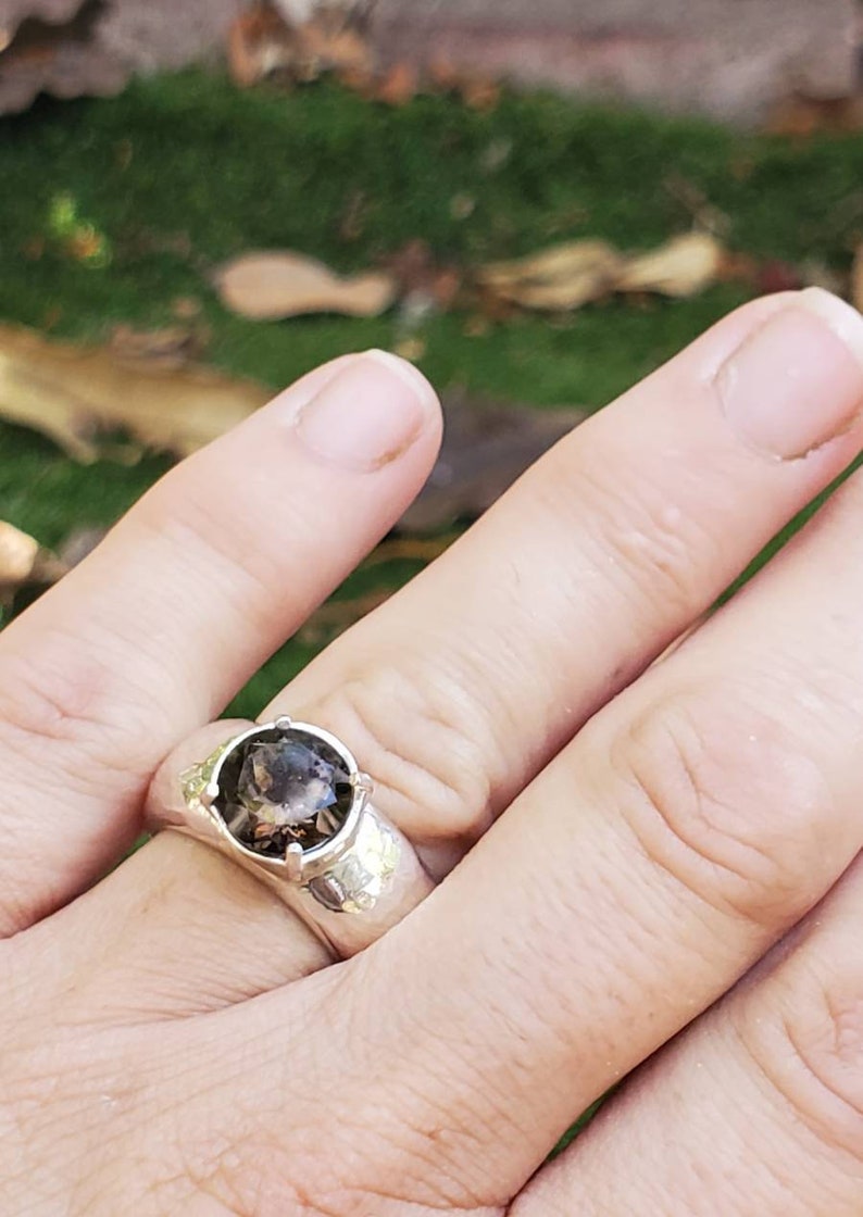 May include: A silver ring with a large, round, smoky quartz gemstone. The ring has a textured band and is set against a blurred green background. The gemstone is a translucent brown color.