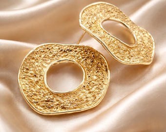 May include: A pair of gold-toned earrings featuring an irregular, organic circular shape with a highly textured, hammered surface. Each earring has a central open cutout and a smooth, polished edge, showcasing a distinct, rugged metallic finish.