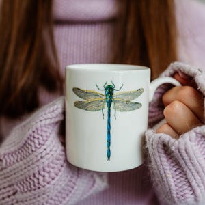 Könnte beinhalten: Weiße Keramiktasse mit einer detaillierten Libellenillustration. Die Libelle hat blaue, grüne und gelbe Akzente. Die Tasse wird von einer Person gehalten, die einen hellvioletten Pullover trägt.