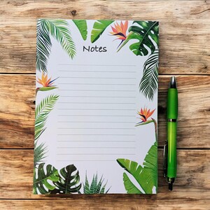 Peut inclure: Bloc-notes ligné avec un motif de feuilles et de fleurs tropicales. Le mot "Notes" est imprimé en haut. Un stylo vert repose sur une surface en bois à côté du bloc-notes.