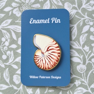 Könnte beinhalten: Eine Emaille-Anstecknadel in Form eines Nautilus-Gehäuses mit cremefarbenen und rotbraunen Streifen sowie einem goldfarbenen Metallrand. Die Anstecknadel ist auf einer blauen rechteckigen Karte mit dem weißen Text Enamel Pin und Willow Paterson Designs befestigt.