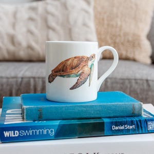 Può includere: Tazza in ceramica bianca con una dettagliata illustrazione di una tartaruga marina nei toni del marrone, verde e blu. La tazza è appoggiata su due libri blu con la scritta "Wild Swimming". La tazza ha un manico curvo.