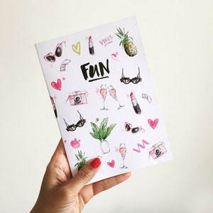 May include: A white notebook with a pink, green and black design. The notebook has the word "FUN" written in black on the front. The design includes hearts, sunglasses, lipsticks, champagne glasses, a camera and a pineapple.