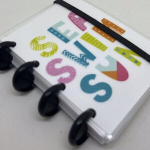 May include: A small, clear plastic notebook with black binding rings and a black elastic band closure. The cover displays the word "SEASONS" in colorful, patterned letters. The notebook is closed and appears to be new.