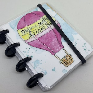 May include: A small notebook with a hot air balloon illustration. The balloon is pink and yellow, with the words "Dreams really do come true." The notebook is held together with black rings and an elastic band.