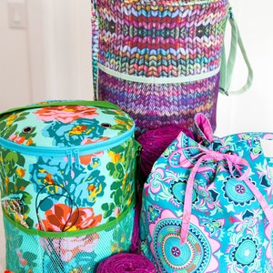May include: Three colorful yarn storage bags with various floral and geometric patterns. One bag is cylindrical with a zipper, another is a drawstring bag, and the third is a tall cylindrical bag with a shoulder strap. Several spools of thread and a ball of yarn are also visible.