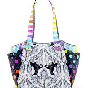 May include: A quilted tote bag featuring a black and white bird and leaf design. The bag has a rainbow striped handle and side pockets with a black polka dot pattern. The bag is a Limited Edition.