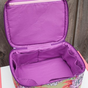 May include: Open, square-shaped cosmetic bag with a floral pattern in shades of green, pink, and purple. The interior is a solid purple, with pockets and a zippered top. The zipper is purple with a pink zipper pull.