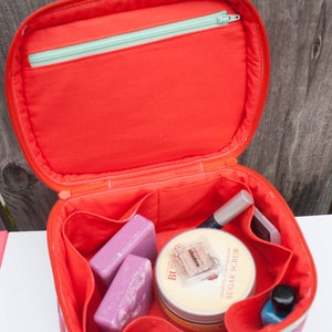 May include: Open red cosmetic bag with a zippered top and multiple compartments. The bag contains beauty products, including two purple bars of soap, a jar of sugar scrub, and nail polish.