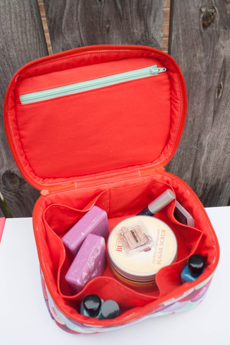 May include: Open red cosmetic bag with a zippered top and multiple compartments. The bag contains beauty products, including two purple bars of soap, a jar of sugar scrub, and nail polish.