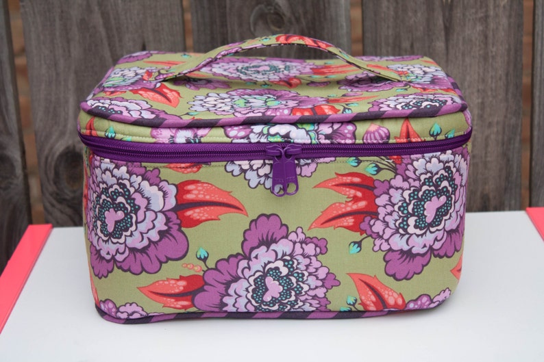 May include: A floral print cosmetic bag with a purple zipper and handle. The bag features a repeating pattern of large purple and pink flowers with red leaves on a light green background. The zipper and handle are a matching purple color.