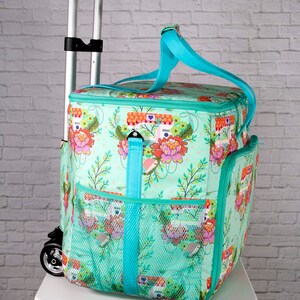May include: A turquoise rolling cooler bag with a floral and camper pattern. The bag has a teal handle and trim, mesh side pockets, and a telescoping handle. The bag is sitting on a white table.
