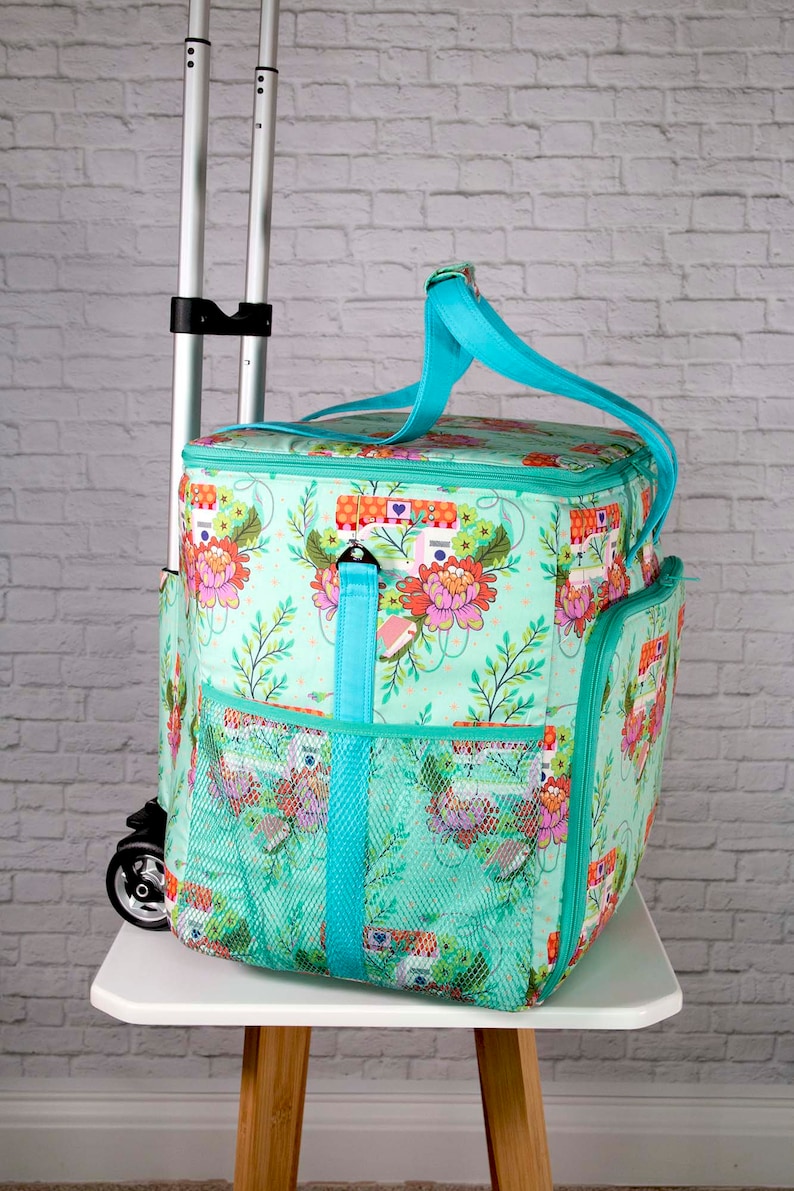 May include: A turquoise rolling cooler bag with a floral and camper pattern. The bag has a teal handle and trim, mesh side pockets, and a telescoping handle. The bag is sitting on a white table.