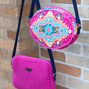 May include: Two crossbody bags with navy blue straps hang against a brick wall. One is oval-shaped, fuchsia with a colorful floral pattern. The other is rectangular, fuchsia with a textured herringbone pattern and a small black label.
