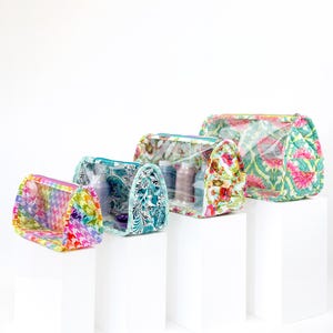 May include: A set of cosmetic bags in various sizes, each with a clear front and patterned fabric. Designs include floral, houndstooth, and botanical prints. The bags are displayed on white pedestals, showcasing their different shapes and sizes.
