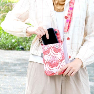 May include: A white and red patterned phone crossbody bag with a matching strap. The bag is held by a person wearing a white shirt, tan pants, and a cream-colored jacket. The strap has a pink and orange design.