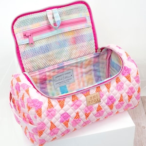 Puede incluir: Una bolsa de viaje acolchada rosa con un patrón repetido de conos de helado rosas sobre un fondo rosa claro. La bolsa tiene una cremallera en la parte superior y un interior colorido con un bolsillo de malla. La bolsa está abierta, revelando el interior.