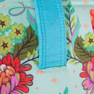 May include: Close-up of a fabric tote bag with a floral pattern in shades of teal, red, orange, and purple. The bag has a bright turquoise handle. The floral design includes flowers, leaves, and other decorative elements.