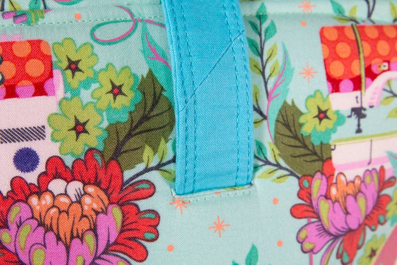 May include: Close-up of a fabric tote bag with a floral pattern in shades of teal, red, orange, and purple. The bag has a bright turquoise handle. The floral design includes flowers, leaves, and other decorative elements.