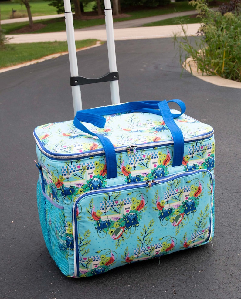 May include: A rolling cooler bag with a blue floral pattern. The bag has a light blue background with a repeating pattern of flowers, houses, and other decorative elements. It features a blue handle, a mesh side pocket, and a telescoping handle.