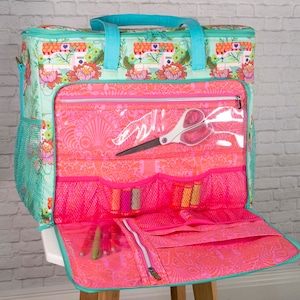 May include: A teal and pink sewing machine themed tote bag with multiple pockets and compartments. The bag features a floral pattern and teal handles. Inside, there are spools of thread, scissors, and pens. The bag is open, revealing its contents.