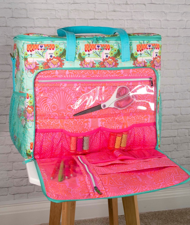 May include: A teal and pink sewing machine themed tote bag with multiple pockets and compartments. The bag features a floral pattern and teal handles. Inside, there are spools of thread, scissors, and pens. The bag is open, revealing its contents.
