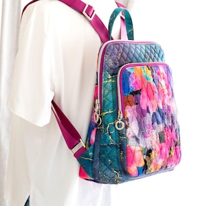 Könnte beinhalten: Ein gesteppter Rucksack mit einem lebendigen Blumenmuster in Rosa, Lila und Blau. Der Rucksack hat ein teal- und goldfarbenes geometrisches Design an den Seiten und einen magentafarbenen Riemen. Er verfügt über silberne Reißverschlüsse und einen oberen Griff.