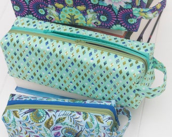May include: Three colorful zippered pencil cases with floral and geometric patterns. The cases have teal, blue, and purple hues, with matching zippers and handles. Three colorful markers are in the foreground.
