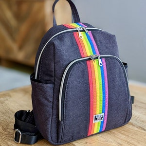 Puede incluir: Mochila de mezclilla gris oscuro con una franja central de arcoíris. La mochila tiene un asa superior, dos compartimentos con cremallera y bolsillos laterales. El texto "BED HOG" es visible en la franja del arcoíris.