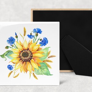 May include: A white ceramic tile with a watercolor painting of a sunflower with yellow petals and a brown center. The sunflower is surrounded by blue cornflowers and wheat stalks.