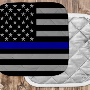 May include: A black and white American flag with a blue stripe across the middle. The flag is on a white quilted pot holder.