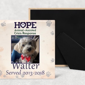 May include: A framed photo featuring a small white dog wearing a red bow tie. The frame has paw prints and the words "HOPE Animal-Assisted Crisis Response" and "Walter Served 2013-2018". The frame is beige with a black backing.
