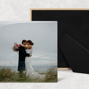 May include: A wedding photo of a couple embracing in a field of tall grass. The bride is wearing a white wedding dress and the groom is wearing a black suit with a kilt. The couple is standing in front of a body of water with a cloudy sky in the background.