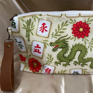 May include: A cream-colored fabric zipper pouch featuring a repeating pattern of green dragons, red flowers, bamboo stalks, and Mahjong tiles with Chinese characters. The pouch includes a brown leather wrist strap and a green tassel charm.