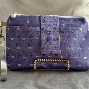 May include: A lavender clutch with a silver wrist strap and a gold-colored metal handle. The clutch features a floral pattern with gold accents and a purple button closure. A white zipper and a pearl-like bead adorn the top.
