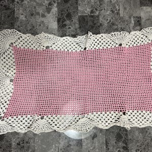 May include: A rectangular, handmade crochet doily with a pink center and a white, scalloped border. The open weave design is visible throughout the piece, showcasing the intricate craftsmanship. This decorative item is suitable for various home decor uses.